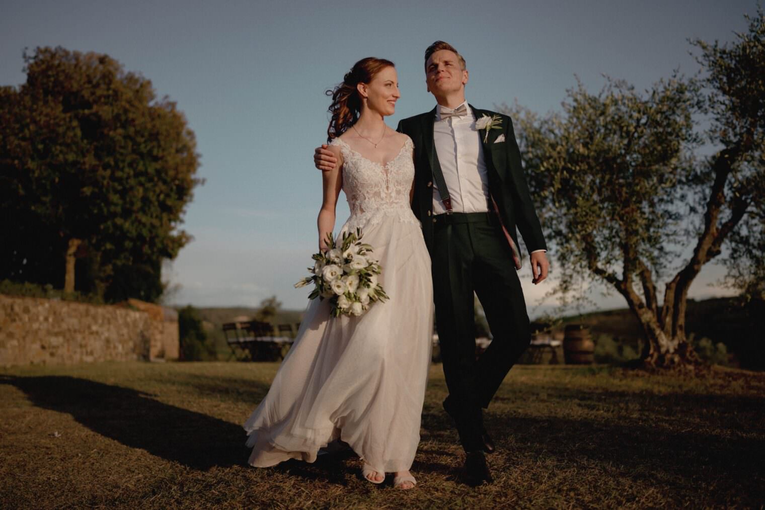 A bride and groom joyfully walk arm in arm outdoors on the lush grass of Fattoria e Villa di Rignana in Tuscany. The bride wears a white lace gown, holding a bouquet of white flowers, while the groom dons a dark suit with a boutonniere. Theyre framed by sunlit trees and a rustic stone wall. Tuscan Wedding Photographer