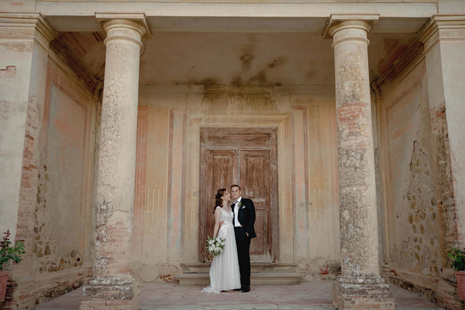 The bride and groom stand in front of the rustic, aged charm of Fattoria e Villa di Rignana in Tuscany. Her white gown contrasts his black tuxedo as they gaze tenderly at each other, symmetrically framed by ornate columns and a weathered door, capturing the villas timeless romance. Tuscan Wedding Photographer