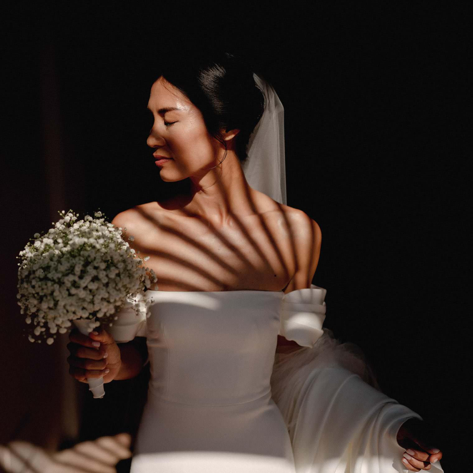 A bride in an off-shoulder white gown and veil stands in soft light, casting striped shadows on her skin. She holds a bouquet of small white flowers, gazing left with a serene expression, as if savoring the moment Federico Pannacci beautifully captures. The dark background accentuates the light on her face and dress. Tuscan Wedding Photographer