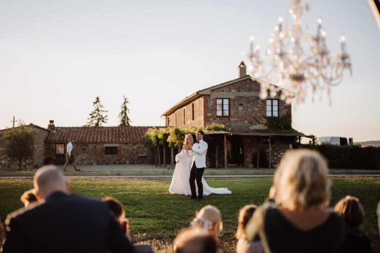 A bride in a white gown and groom in a white jacket share their first dance on a grassy lawn at one of the top wedding venues in Tuscany, set against a rustic stone house at sunset. A chandelier hangs nearby, as guests watch from their seats, with golden sunlight casting a warm and romantic glow on the couple. Tuscan Wedding Photographer
