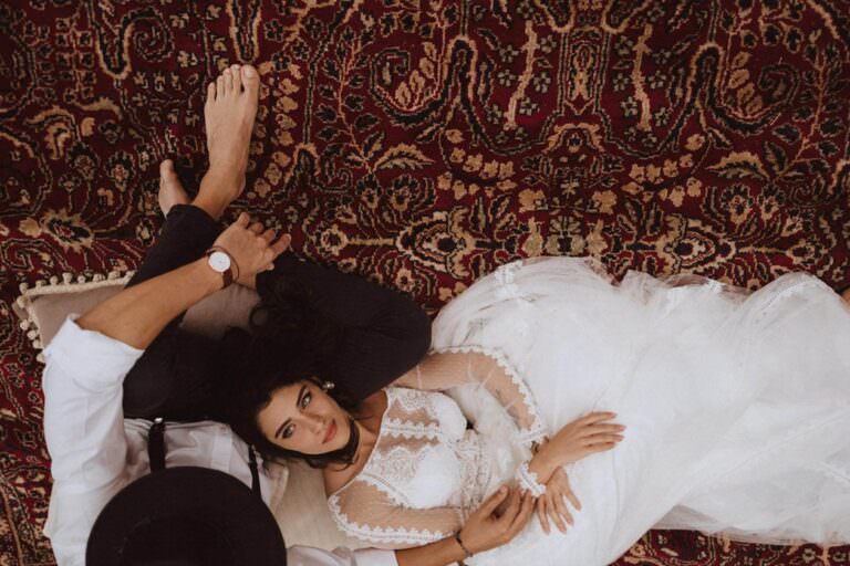 A bride in a lace wedding dress with long sleeves lies on a red patterned rug, her head on her partners lap, dreaming of how to plan your elopement in Italy. Her dark hair and red lipstick complement his white shirt and black hat. Both appear relaxed and content, embodying loves serene spontaneity. Tuscan Wedding Photographer