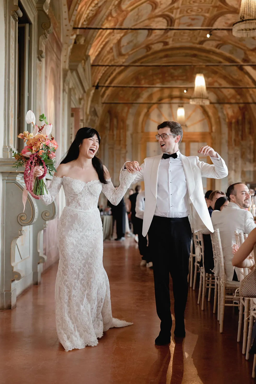 A joyful bride in a lace off-shoulder gown and groom in a white tuxedo walk hand in hand down an ornate hall, celebrating their big day. Capture moments like these when you know how to find the perfect destination wedding photographer. Tuscan Wedding Photographer