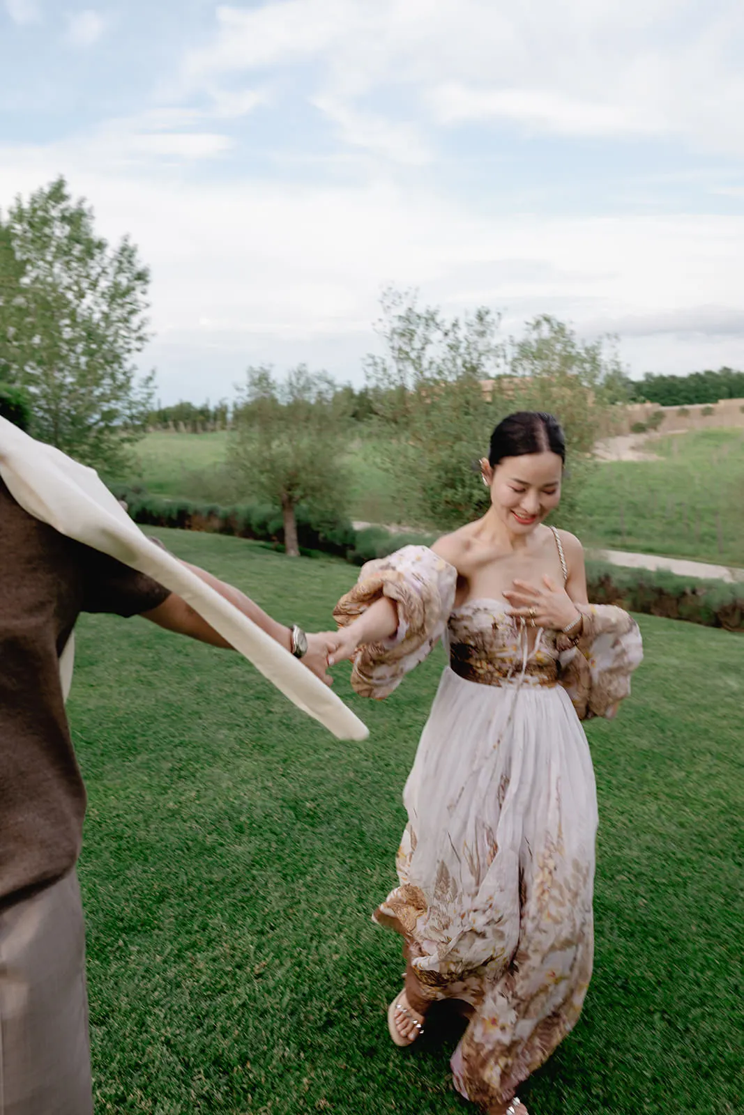 A woman in a flowing, off-shoulder dress with puffed sleeves smiles while holding hands with a person partly out of frame on a lush green lawn—capturing the joy you want when learning how to find the perfect destination wedding photographer. Tuscan Wedding Photographer