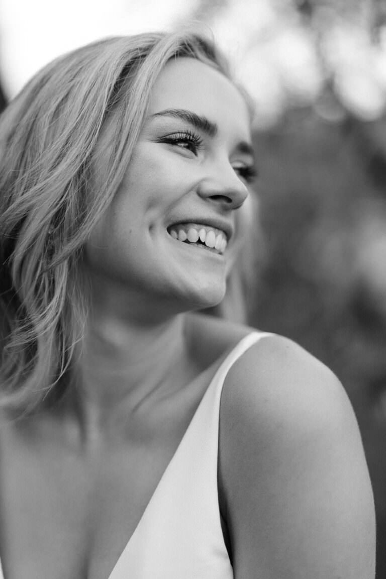 Black and white photo of a woman with shoulder-length blonde hair smiling and looking to the side, reminiscent of joyful moments at an intimate wedding at Villa Petrea in Tuscany. She is wearing a sleeveless top against a softly blurred background, highlighting her joyful expression and dimples. Tuscan Wedding Photographer