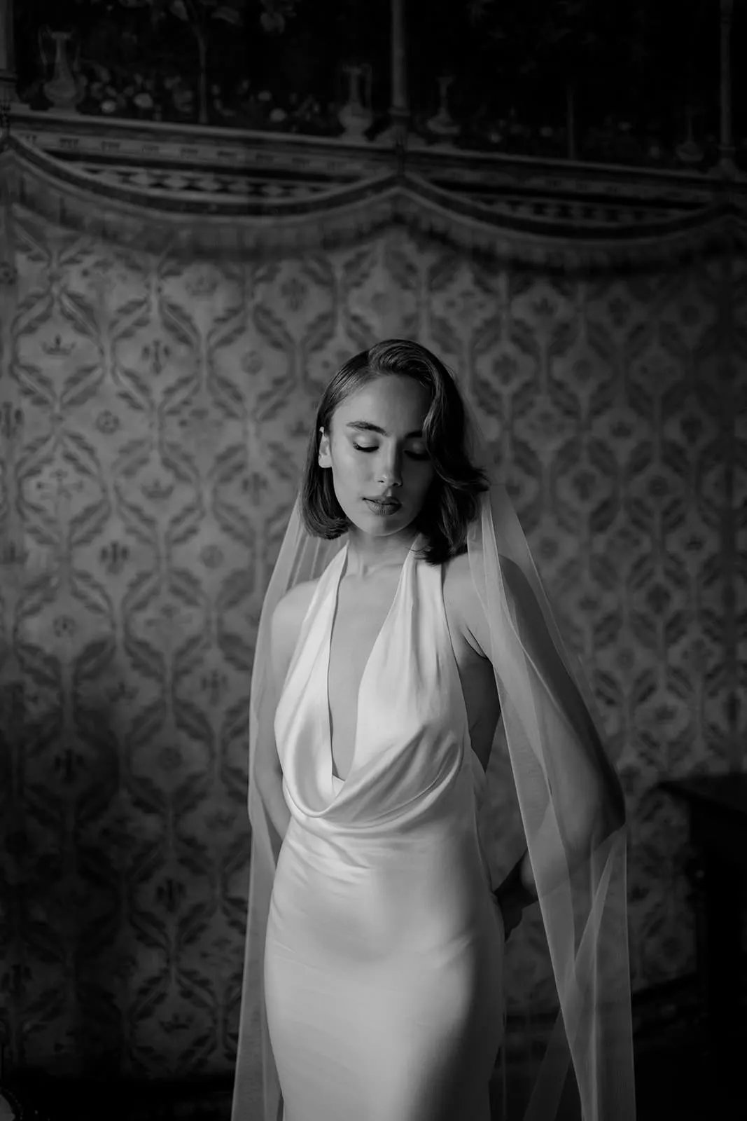 A woman in a silky, sleeveless, deep V-neck gown and long veil stands with eyes closed against ornate wallpaper. This black and white image evokes timeless elegance—capturing the serenity you'd want when learning how to find the perfect destination wedding photographer. Tuscan Wedding Photographer