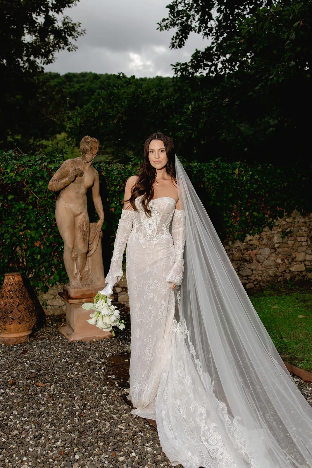 A bride in a long, off-the-shoulder lace wedding gown with sheer sleeves stands outdoors on gravel, holding white flowers—captured beautifully as if by someone who knows how to find the perfect destination wedding photographer. Tuscan Wedding Photographer