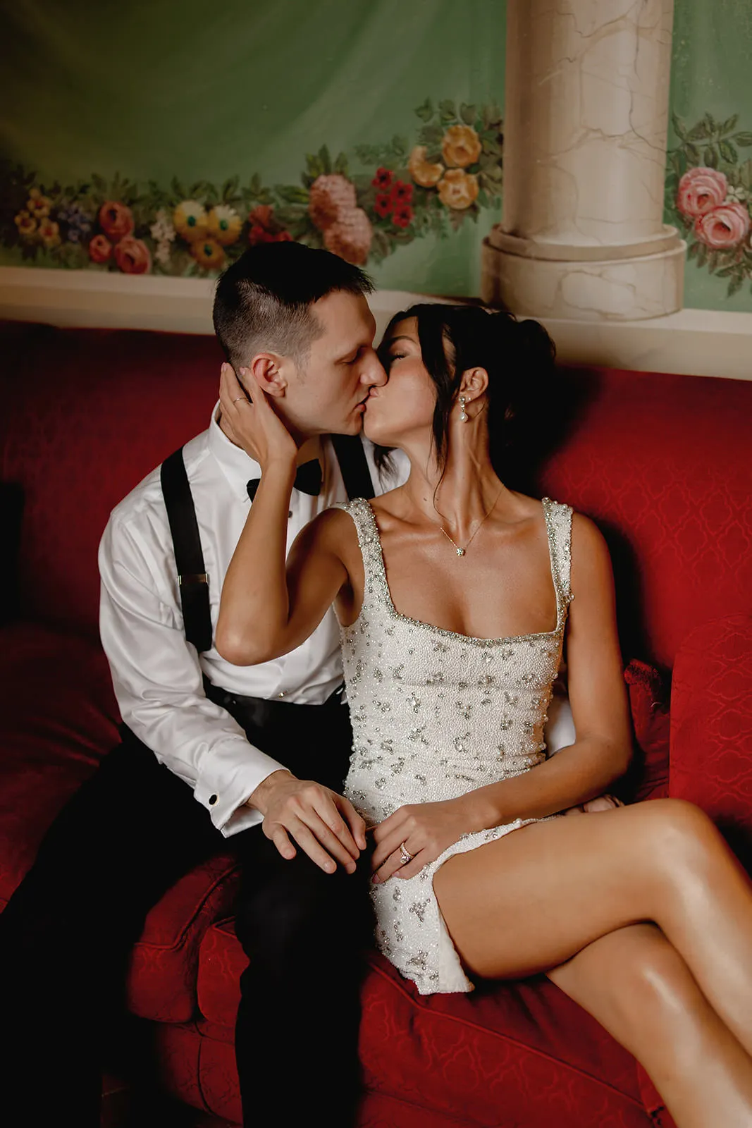A couple sits on a red couch, kissing—capturing the magic you’d want from your destination wedding photos. The man wears a white shirt with suspenders; the woman, a sleeveless embellished dress. Behind them is a floral mural—a scene worthy of the perfect destination wedding photographer. Tuscan Wedding Photographer