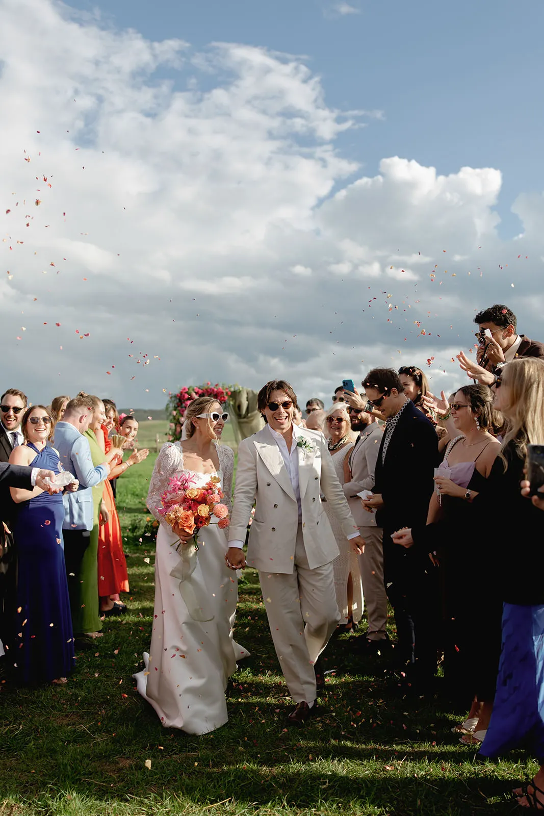 A newlywed couple in white suits strolls hand in hand down an outdoor aisle as guests throw flower petals, celebrating under a bright sky. Capturing moments like this is why choosing how to find the perfect destination wedding photographer matters. Tuscan Wedding Photographer