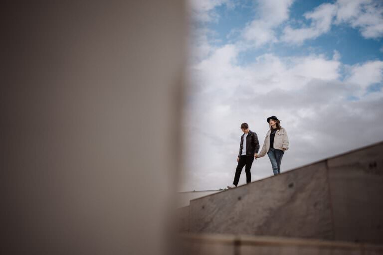Two people walking hand in hand on a slanted concrete surface, perfect inspiration for what to wear during your engagement shoot. Outdoors under a partly cloudy sky, one sports a black jacket and pants, the other a white jacket and jeans. The blurred foreground adds artistic depth to the image. Tuscan Wedding Photographer