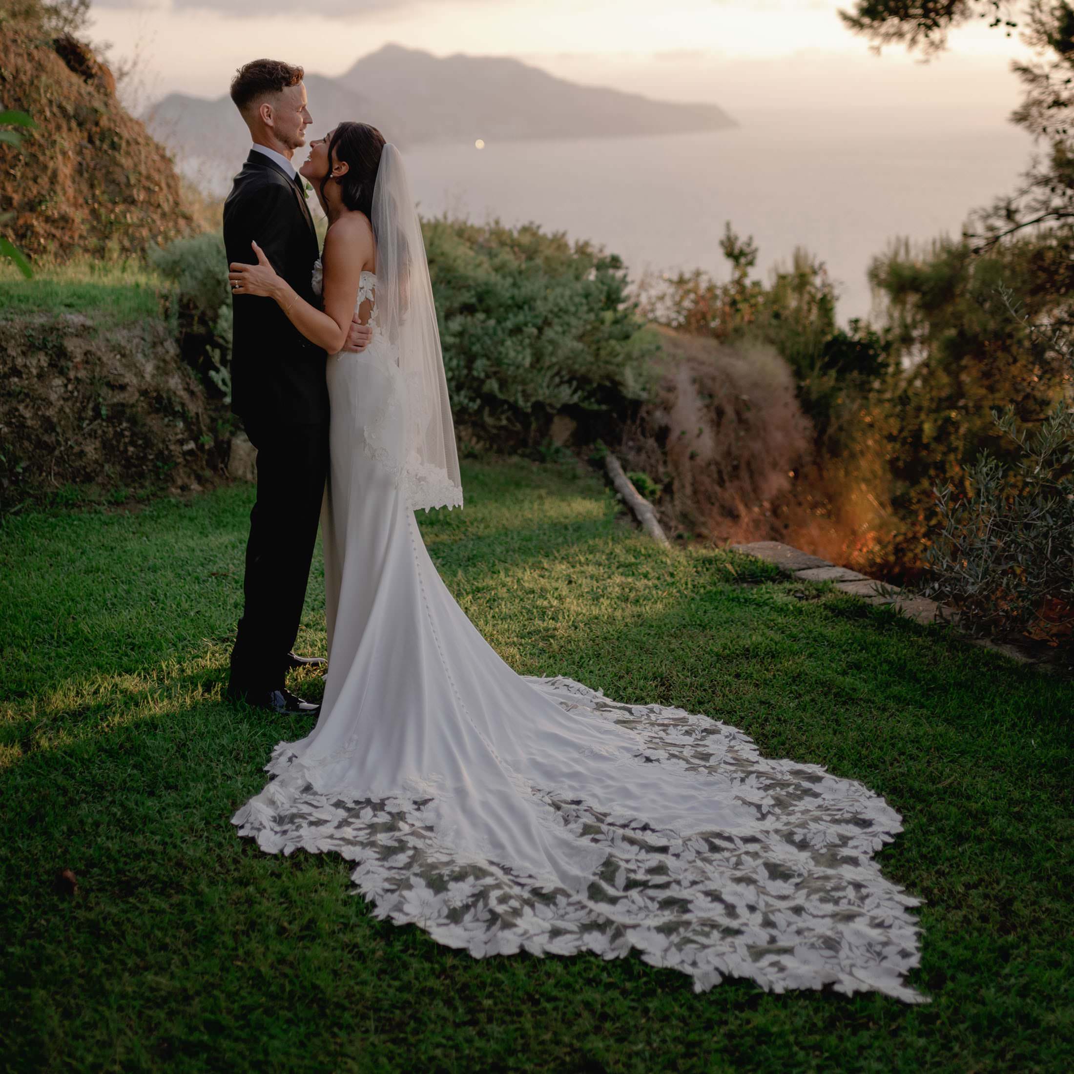 Bride and groom embrace on a grassy hill overlooking the scenic, hazy expanse of the Amalfi Coast. The brides long white gown with intricate lace details trails behind her at their wedding at Relais Blu. Sunlight filters through trees, casting a warm glow on the couple and landscape. Tuscan Wedding Photographer