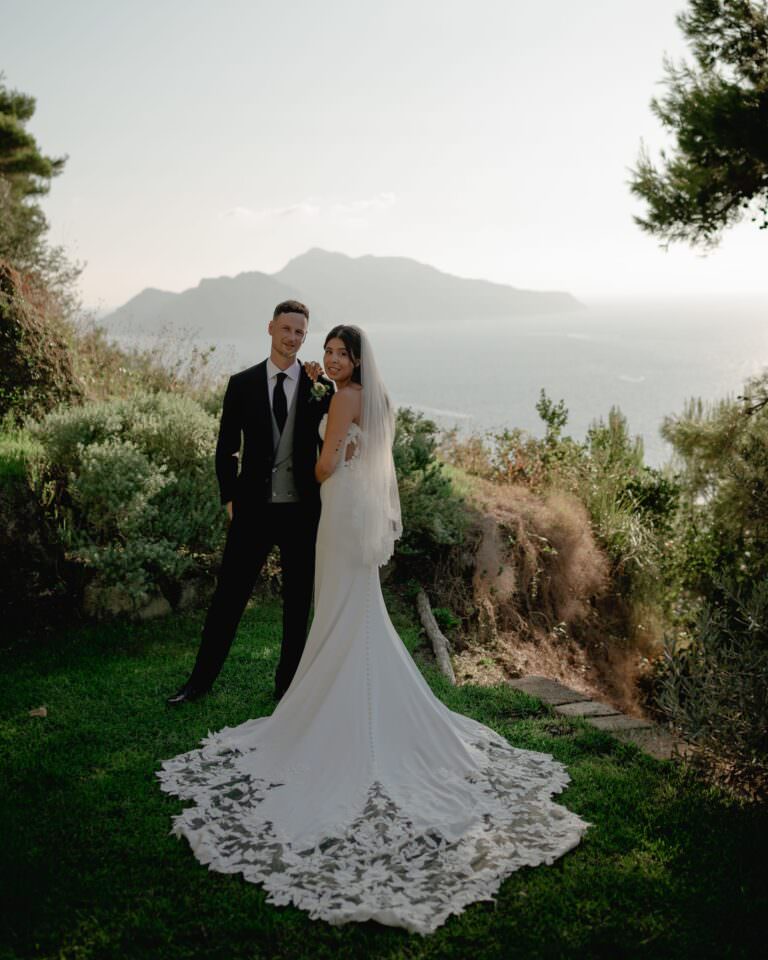 A bride and groom stand on a grassy cliff along the Amalfi Coast with the ocean and an island in the background. The bride wears a long, lace-trimmed white gown and veil, while the groom is in a black suit. The scene at Relais Blu is surrounded by lush greenery and bathed in soft, natural light. Tuscan Wedding Photographer