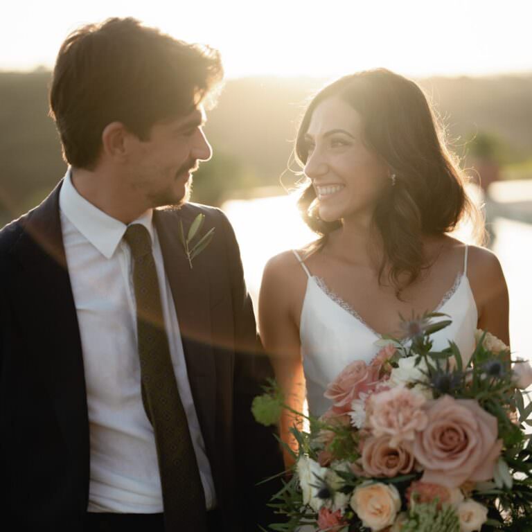 A bride and groom, glowing in the soft, golden sunlight of an elegant wedding at Tenuta di Monaciano in Tuscany, smile at each other. The bride holds a bouquet with pink and white flowers, wearing a white dress with delicate straps. The groom wears a dark suit with a white shirt and dark tie against the blurred greenery backdrop. Tuscan Wedding Photographer