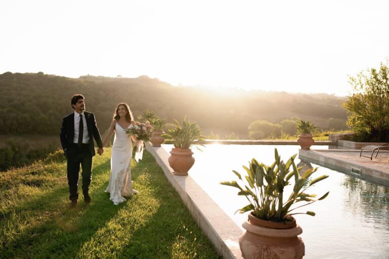A couple epitomizes timeless elegance as they stroll hand-in-hand by a pool at sunset during their wedding at Tenuta di Monaciano. The woman, in her white dress with a bouquet, and the man, in his black suit, are bathed in the suns warm glow against rolling hills and lush potted plants. Tuscan Wedding Photographer