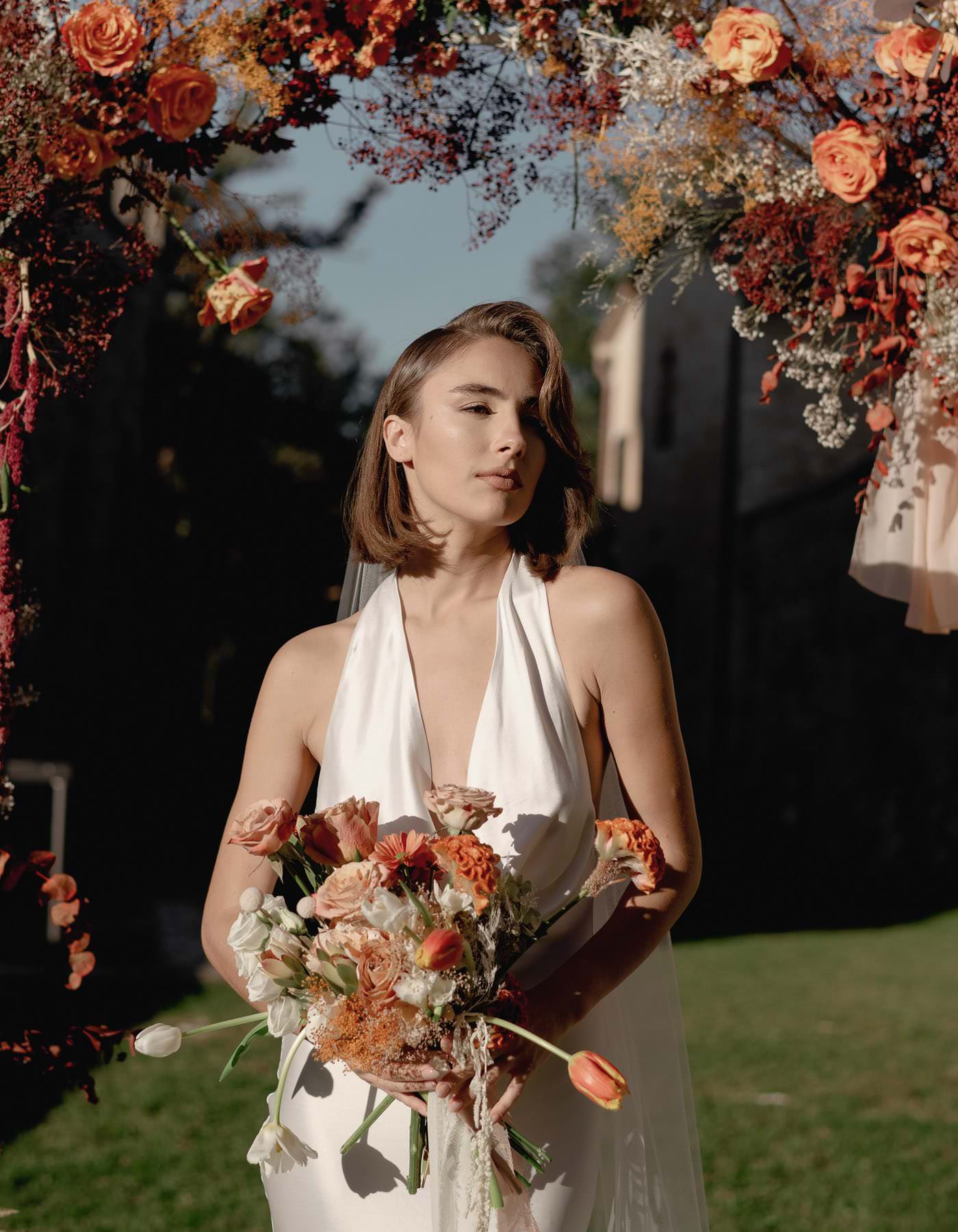 A bride in a sleeveless white gown holds an autumn-themed bouquet of orange and white flowers, standing under a floral arch adorned with vibrant blooms. The sunlit path, framed by greenery at Palazzo Al Piano, provides a serene outdoor setting for this enchanting wedding. Tuscan Wedding Photographer