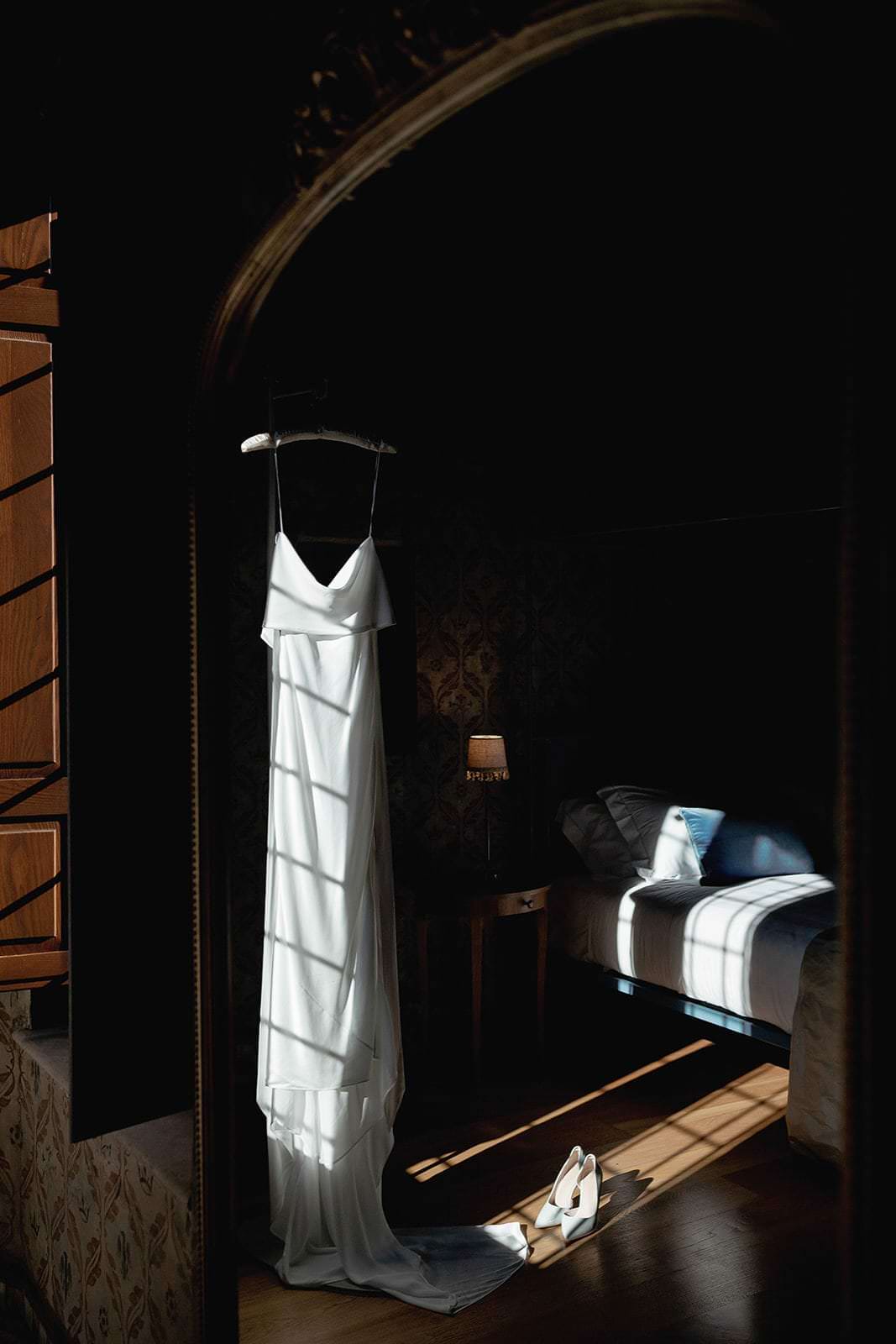 A Grace Loves Lace Suki dress elegantly hangs on a hanger, its white satin illuminated by sunlight streaming through wooden shutters in a dimly lit room. The dress casts a shadow near white heels on the floor, evoking the enchanting vibe of a wedding in Tuscany. A made bed and small lamp are partially visible nearby. Tuscan Wedding Photographer