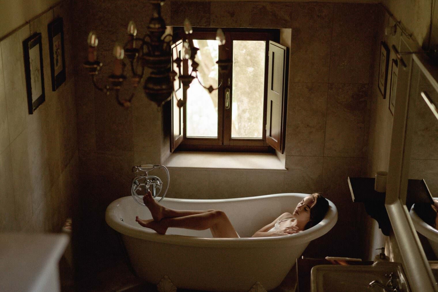A person relaxes like a Grace Loves Lace bride in a clawfoot bathtub in a dimly lit, rustic bathroom with beige tiled walls. A window with wooden shutters lets in natural light, while a chandelier dangles above. Framed pictures adorn the walls, and a sink with a mirror is partially visible on the right. Tuscan Wedding Photographer