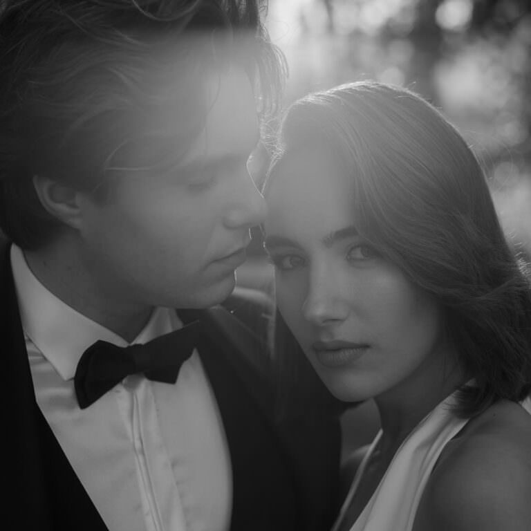 A black-and-white photo of a couple in formal attire captures the timeless elegance of their Palazzo Al Piano wedding. The man, in a suit and bow tie, leans gently towards the woman, who gazes calmly at the camera. Soft, natural lighting outdoors heightens the intimate mood. Tuscan Wedding Photographer