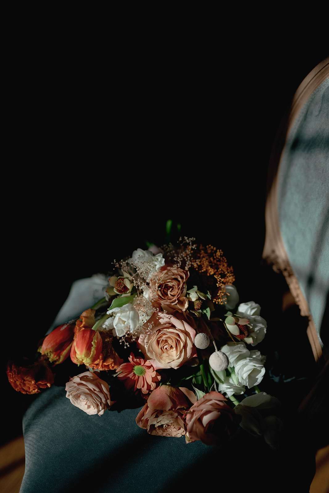 A bouquet of roses and tulips in shades of pink, red, and white graces a teal velvet chair. Captured by Federico Pannacci Photography at a Palazzo Al Piano wedding in Tuscany, the flowers’ vibrant colors and delicate textures are beautifully highlighted against the dark background. Tuscan Wedding Photographer