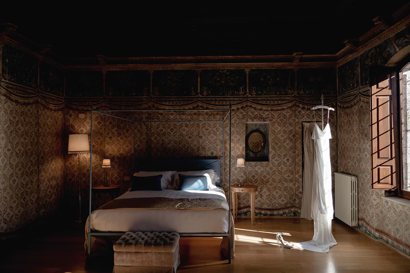 A dimly lit bedroom in a classical style, reminiscent of a romantic Palazzo al Piano wedding location in Tuscany. A white dress hangs elegantly near the open window, where sunlight streams onto the four-poster bed. The room exudes timeless charm with its patterned wallpaper and cushioned ottoman. Tuscan Wedding Photographer