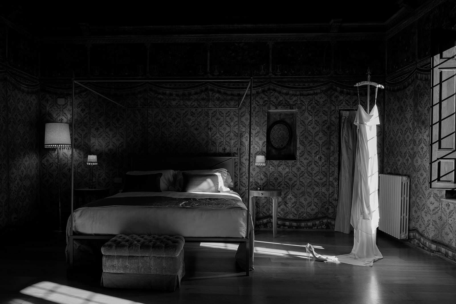 A dimly lit bedroom, reminiscent of a Palazzo al Piano wedding location in Tuscany, features a four-poster bed with pillows and a blanket, flanked by two lamps on tables. A cushioned bench rests at the foot. A long, elegant dress hangs by the window, with sunlight streaming in across patterned walls and wooden floors. Tuscan Wedding Photographer