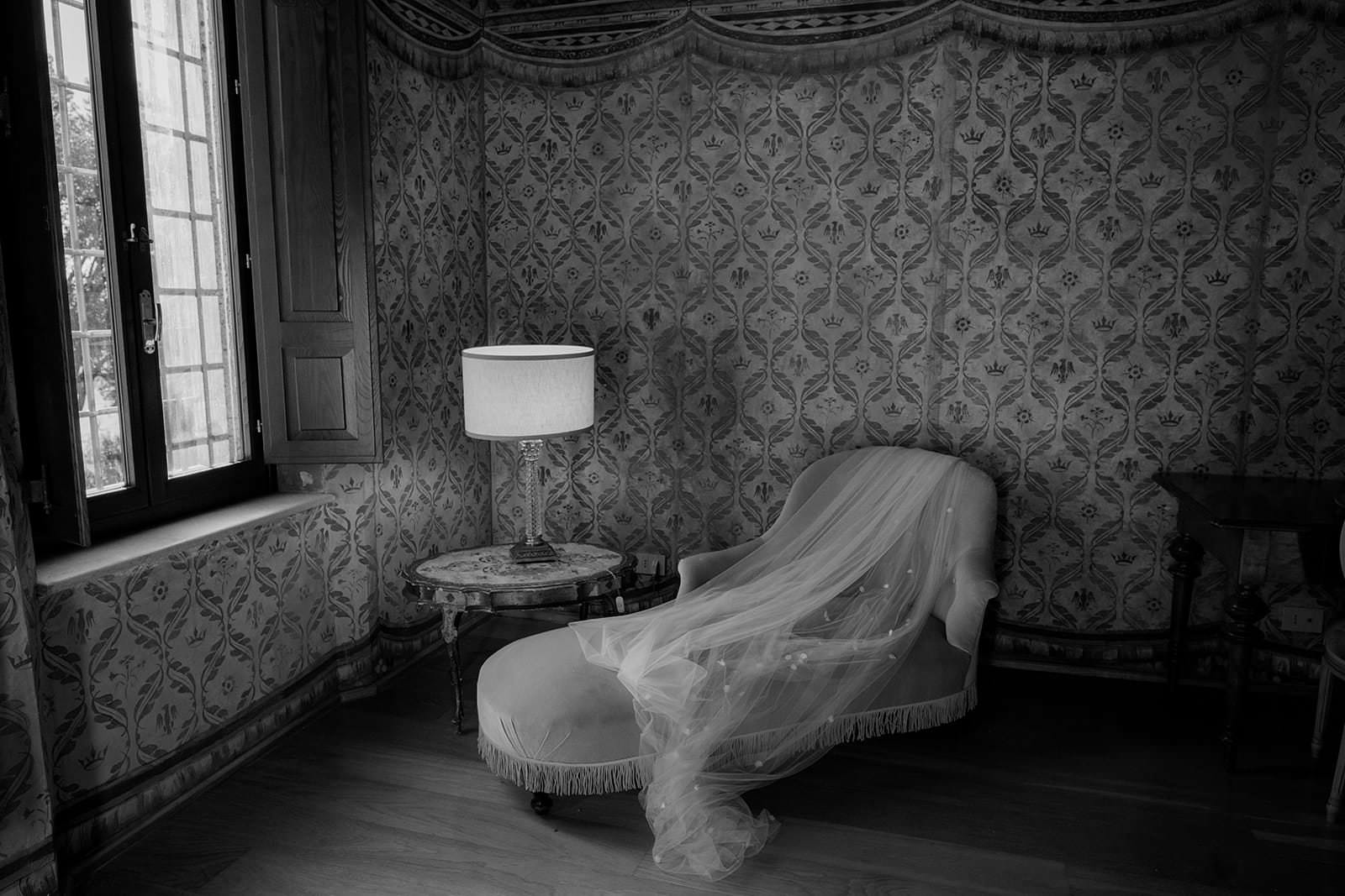 A vintage room with patterned wallpaper and wooden floors exudes a serene and nostalgic ambiance, reminiscent of a Palazzo al Piano wedding location in Tuscany. A chaise lounge is draped with a flowing, translucent veil beside a round table holding a lit lamp beneath elegant curtains. Tuscan Wedding Photographer