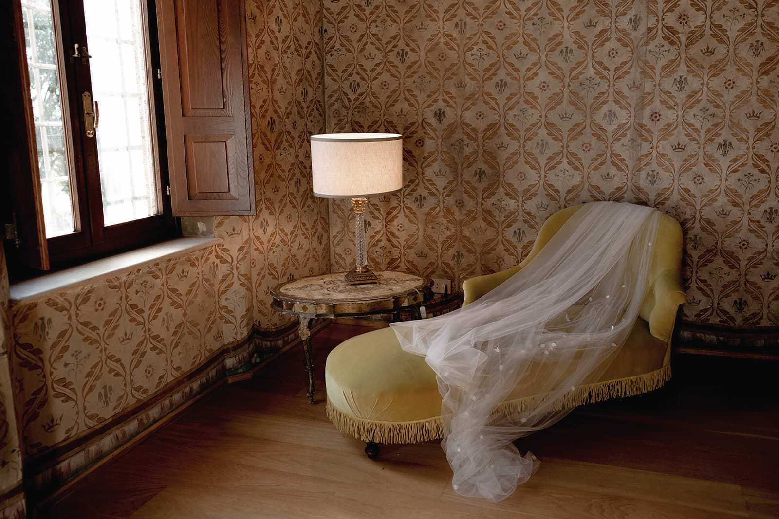 A room with vintage decor at the Palazzo al Piano Wedding Location in Tuscany features a yellow chaise lounge draped with a sheer white veil. A round wooden side table holds a lit lamp with a beige shade. The walls and window curtains display intricate floral patterns, and sunlight filters through. Tuscan Wedding Photographer