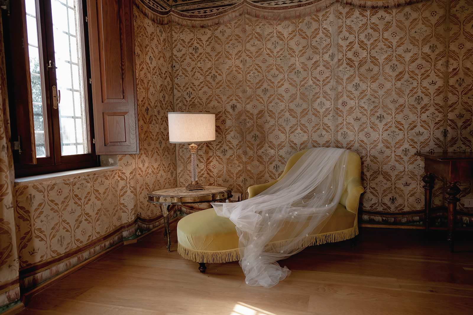 A luxurious room in the Palazzo al Piano, a renowned wedding location in Tuscany, features ornate wallpaper and a yellow chaise lounge draped with sheer white fabric. A round side table with a lamp sits nearby, while sunlight streams through the large window, casting soft light onto the hardwood floor. Tuscan Wedding Photographer