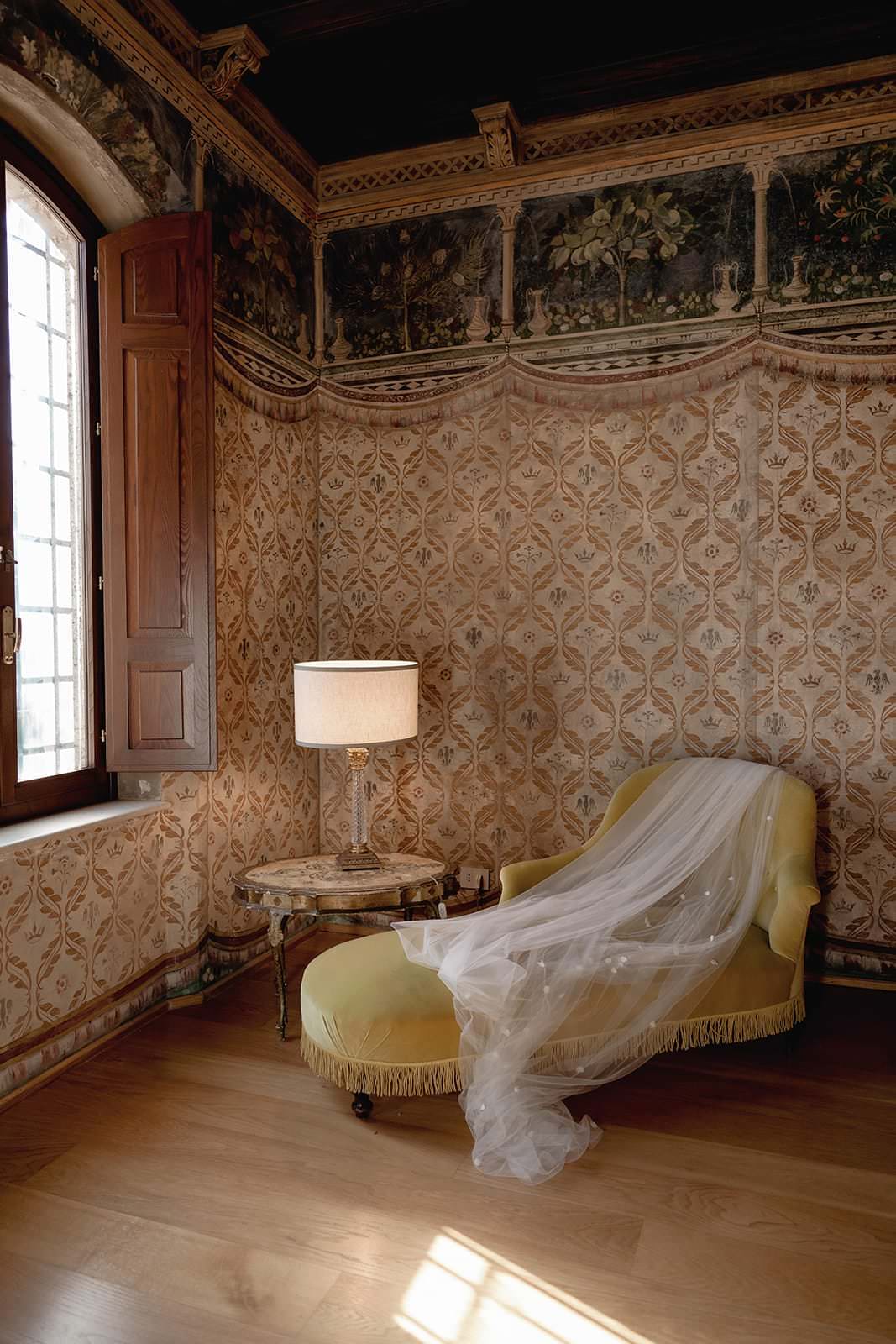 A vintage room with ornate wallpaper and a wooden floor features a yellow chaise lounge draped with a long, flowing white veil. A small round marble table with a lamp sits beside it. Sunlight streams in through the open window from Tuscanys Palazzo al Piano, making it an ideal wedding location. Tuscan Wedding Photographer