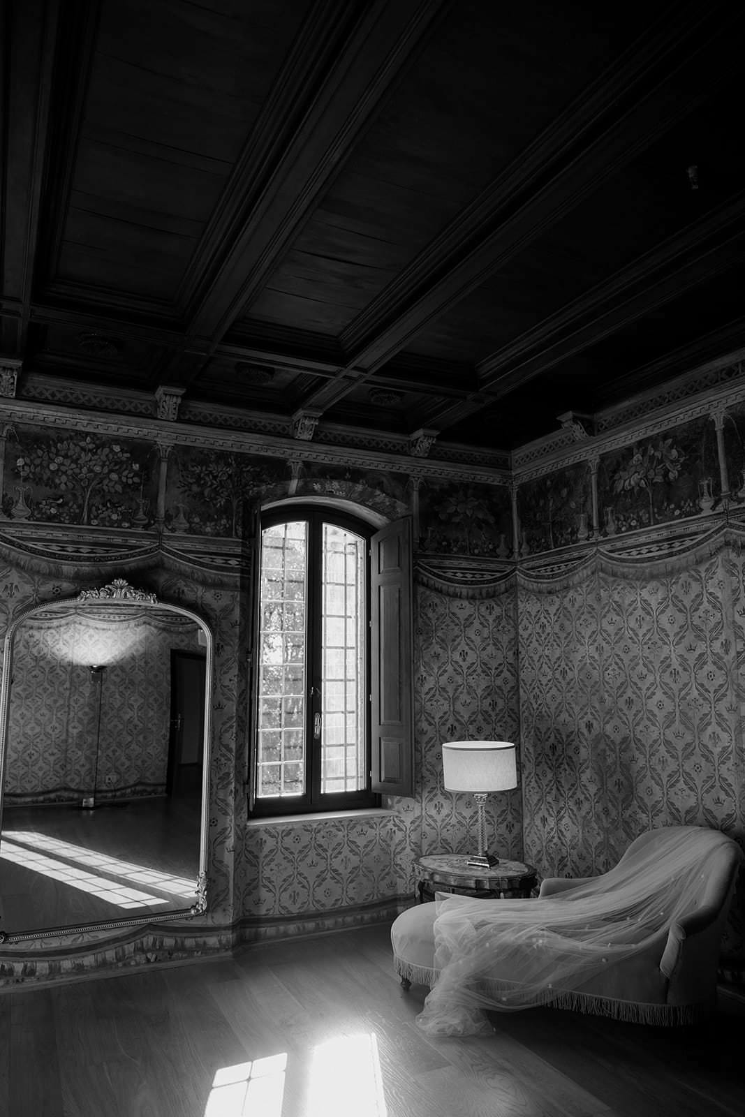A black and white photo captures the ornate elegance of a room at Palazzo al Piano, a stunning wedding location in Tuscany. Intricate wallpaper frames a large window with open shutters, as sunlight dances on a vintage mirror. A tufted chaise lounge beside a lit floor lamp graces the wooden floor. Tuscan Wedding Photographer