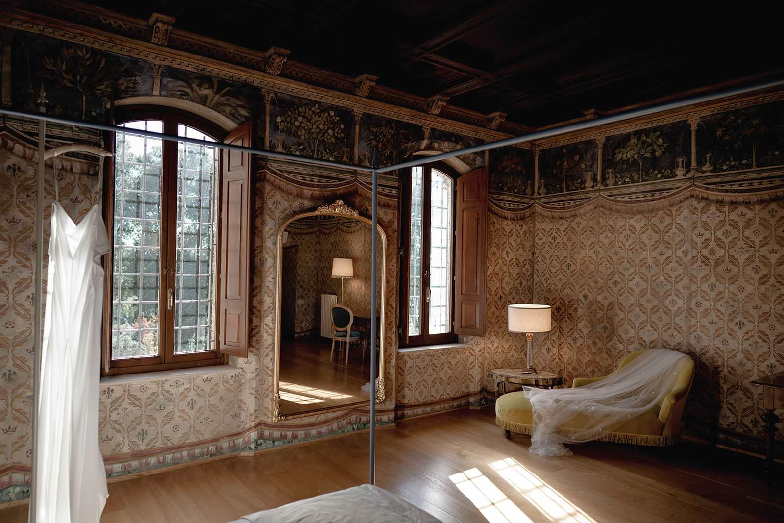 A luxurious vintage room at the Palazzo al Piano wedding location in Tuscany features patterned wallpaper and ornate moldings. A white wedding dress hangs near tall, arched windows with wooden frames. A large mirror reflects a chair and table, while a chaise lounge basks in soft light from another window. Tuscan Wedding Photographer