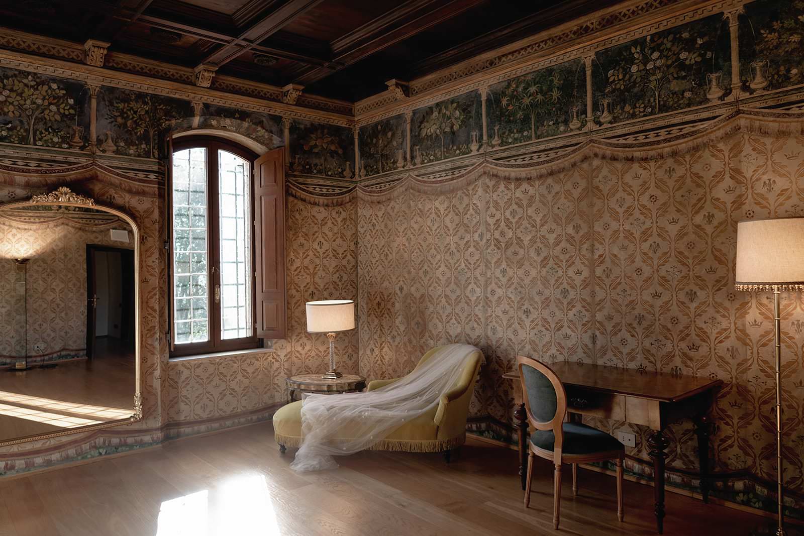 A richly decorated room in the Palazzo al Piano wedding location in Tuscany features ornate floral wallpaper and a large, intricately framed wall mirror. Natural light pours through a window onto a yellow chaise longue with a draped white veil, beside a table with an upholstered chair and a lit floor lamp. The ceiling is wooden. Tuscan Wedding Photographer