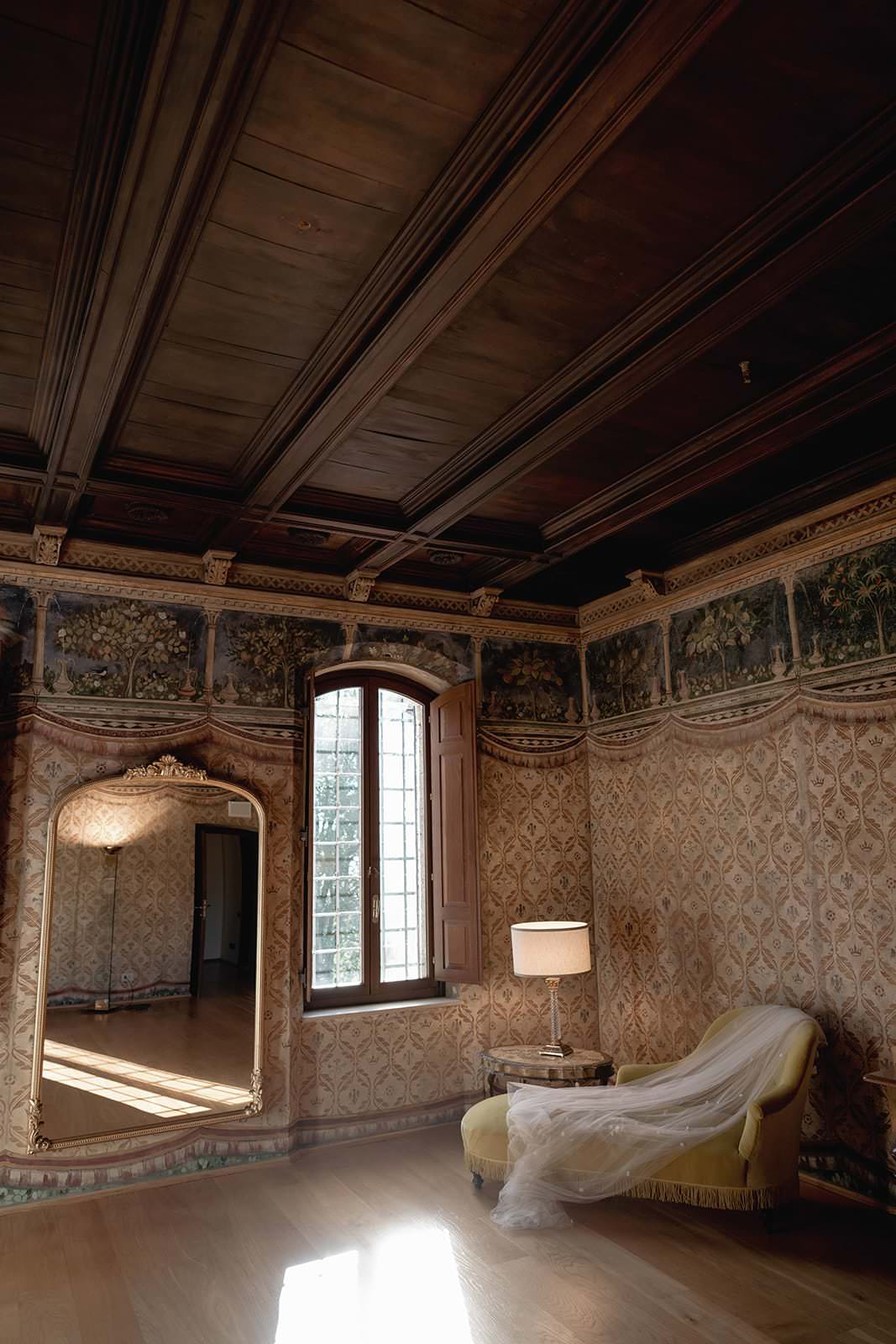 A vintage room at the Palazzo al Piano in Tuscany features ornate wallpaper and a high wooden ceiling. A large mirror reflects the elegance, while wooden shutters filter soft light over a yellow chaise longue draped in sheer white fabric, setting a romantic scene perfect for a wedding backdrop. Tuscan Wedding Photographer