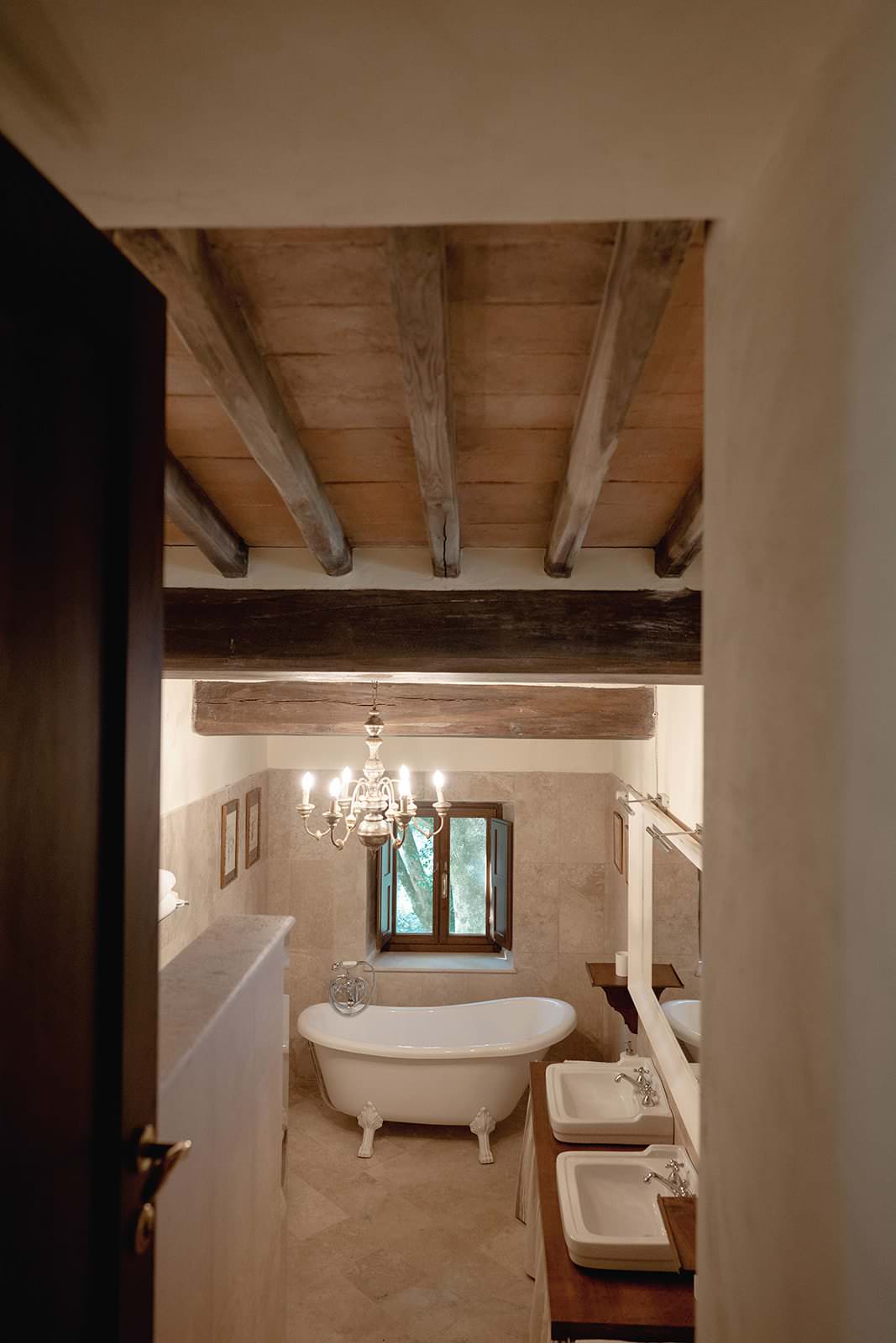 A rustic bathroom in the Palazzo al Piano Wedding Location in Tuscany features a freestanding white bathtub beneath a window. A chandelier hangs from the wooden beam ceiling, while two sinks with mirrors line the right side. The earthy tones of walls and floor create a cozy atmosphere, enhanced by an open door framing the scene. Tuscan Wedding Photographer