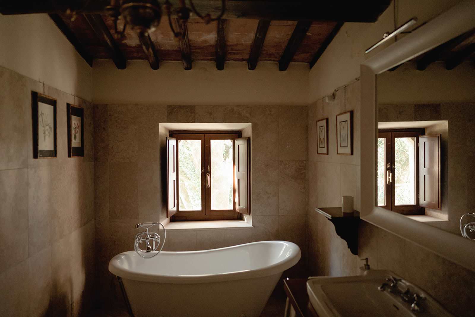 A rustic bathroom, reminiscent of a charming Palazzo al Piano wedding location in Tuscany, features a white freestanding bathtub beneath a wooden-framed window with open shutters. Light brown tiled walls display small artwork, while exposed beams add warmth. A white sink is partially visible below a mirror. Tuscan Wedding Photographer