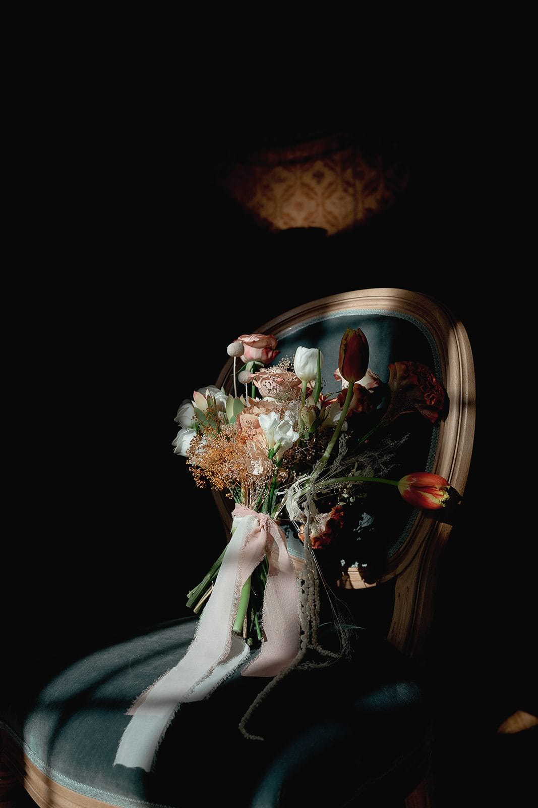 A bouquet of assorted flowers, including tulips and carnations, rests on a vintage-style wooden chair with a light green cushion. In the style of Federico Pannacci Photography, soft natural light highlights the bouquet against a dark background, casting subtle shadows as delicate ribbons hang gracefully. Tuscan Wedding Photographer