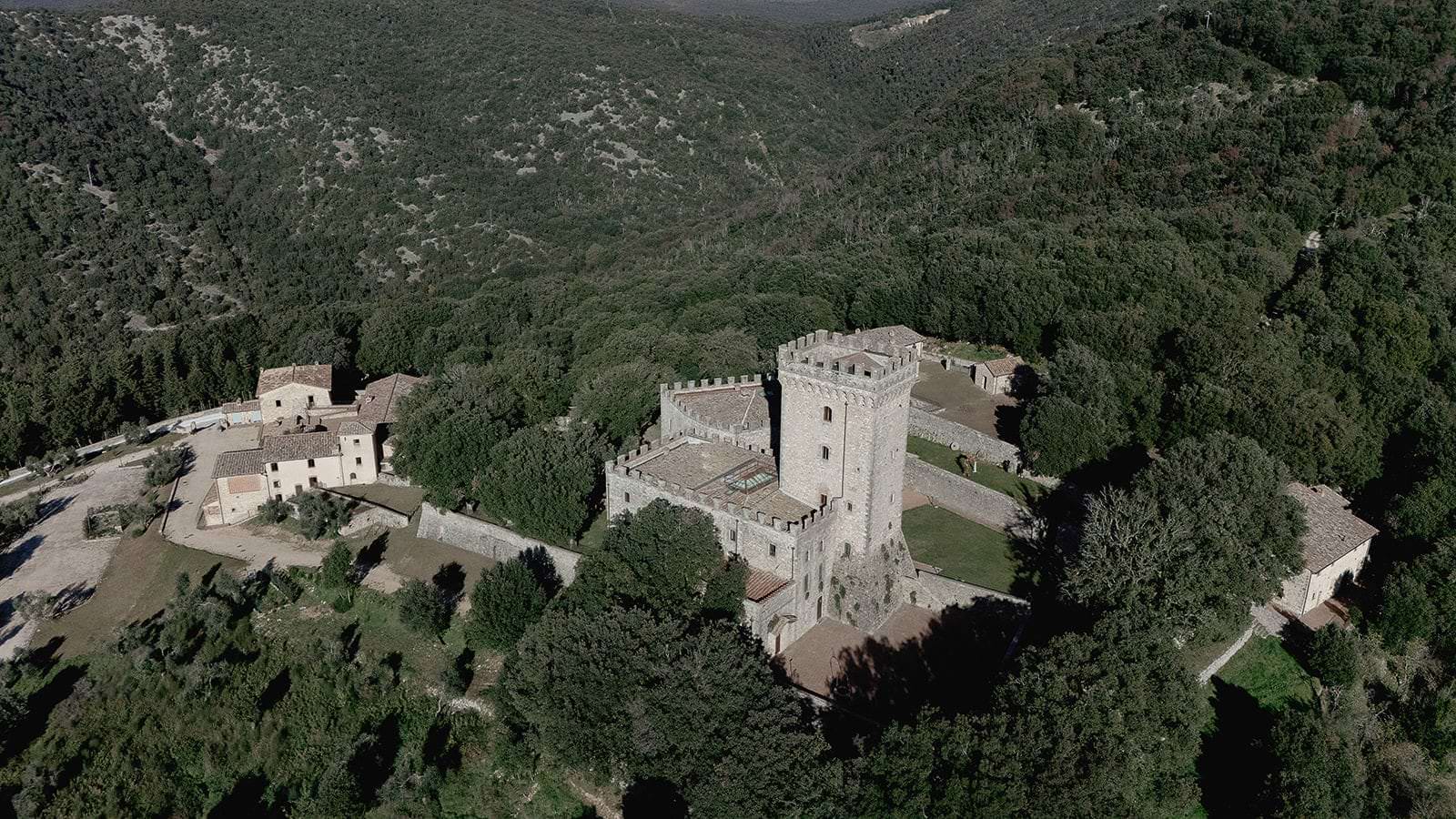 Aerial view of a medieval stone castle in Tuscany surrounded by dense greenery, perfect for an exclusive wedding by Federico Pannacci. The castle boasts a tall central tower, crenellated walls, and adjoining buildings. Nearby rustic houses dot the forested hills, creating a serene and secluded atmosphere. Tuscan Wedding Photographer