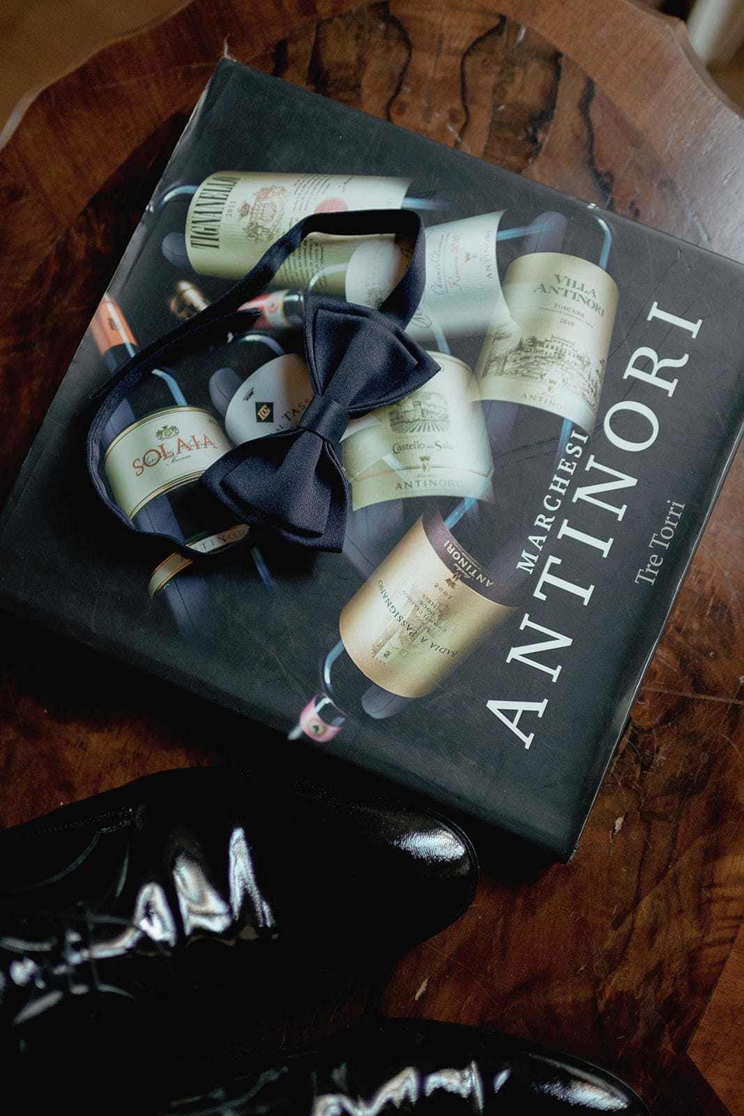 A large box with various wine bottle images and Marchesi Antinori text evokes the charm of an exclusive wedding in Tuscany by Federico Pannacci. A navy bow tie rests elegantly on top, surrounded by a wooden surface, while shiny black dress shoes add a touch of sophistication at the bottom. Tuscan Wedding Photographer