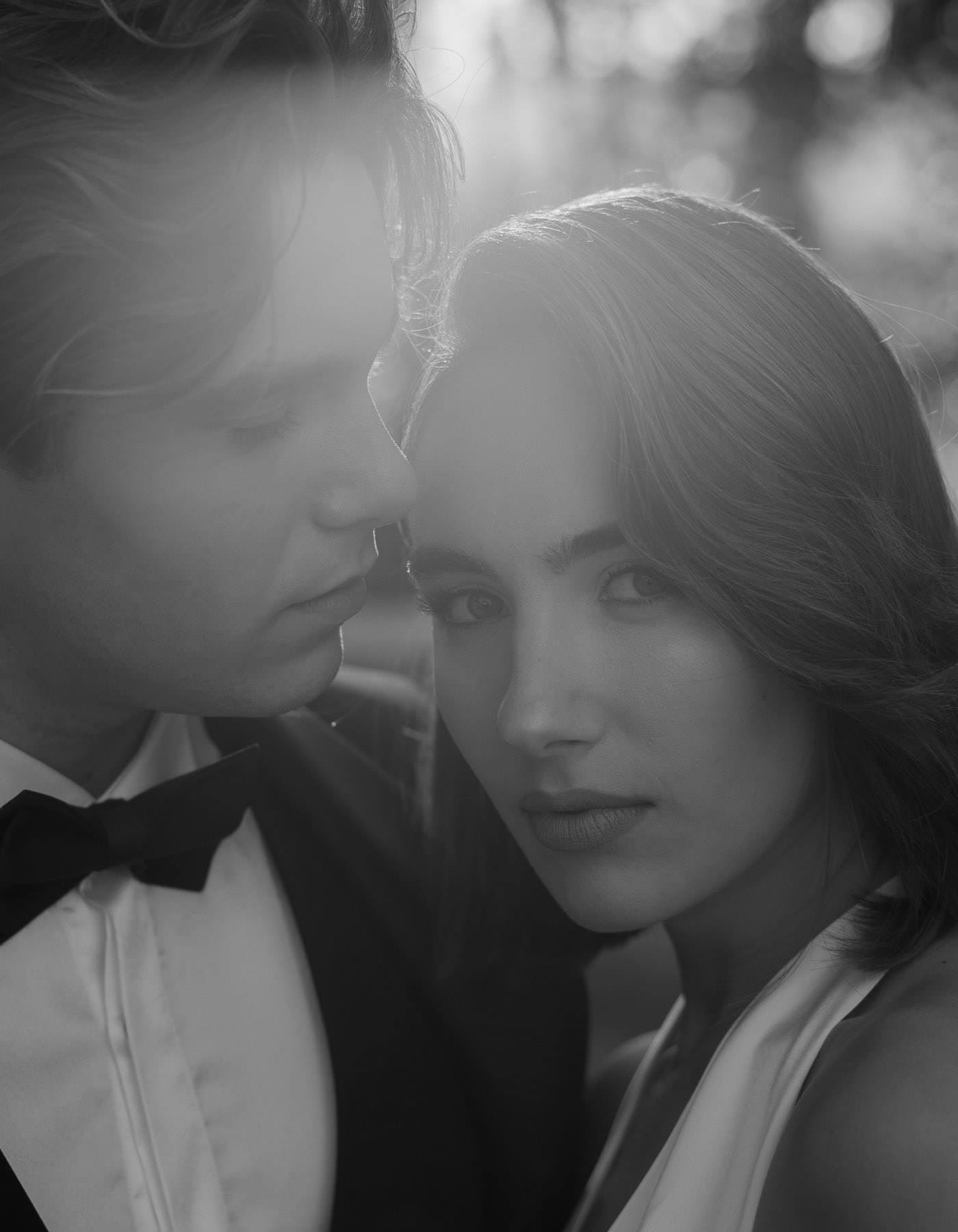 A black-and-white close-up photo captures a couple at a Palazzo Al Piano wedding. The man in a tuxedo and bow tie gazes lovingly at the woman, who looks directly at the camera. Sunlight filters through their hair, creating a soft glow against an abstract background of trees. Tuscan Wedding Photographer