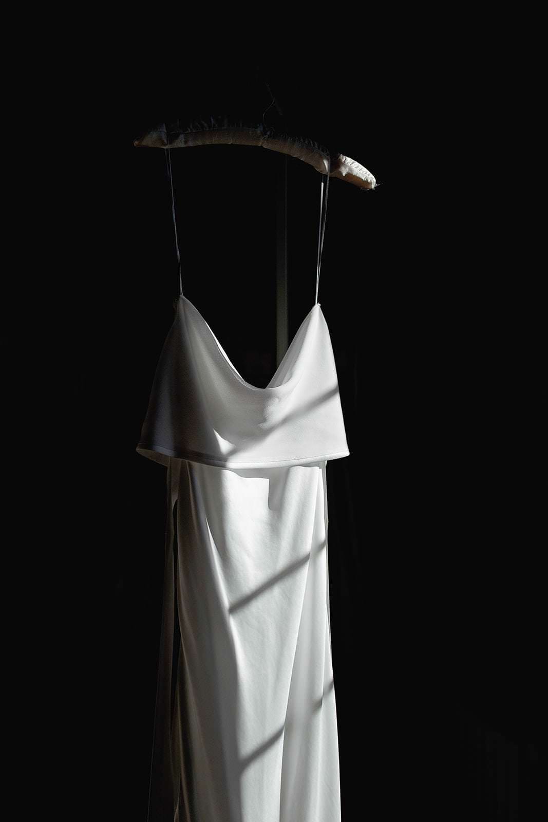A white satin dress with thin straps hangs on a padded hanger against a black background, reminiscent of Federico Pannacci Photography. The dress features a draped cowl neckline and flowing silhouette. Bright, diagonal sunlight highlights its smooth texture, casting soft shadows. Tuscan Wedding Photographer