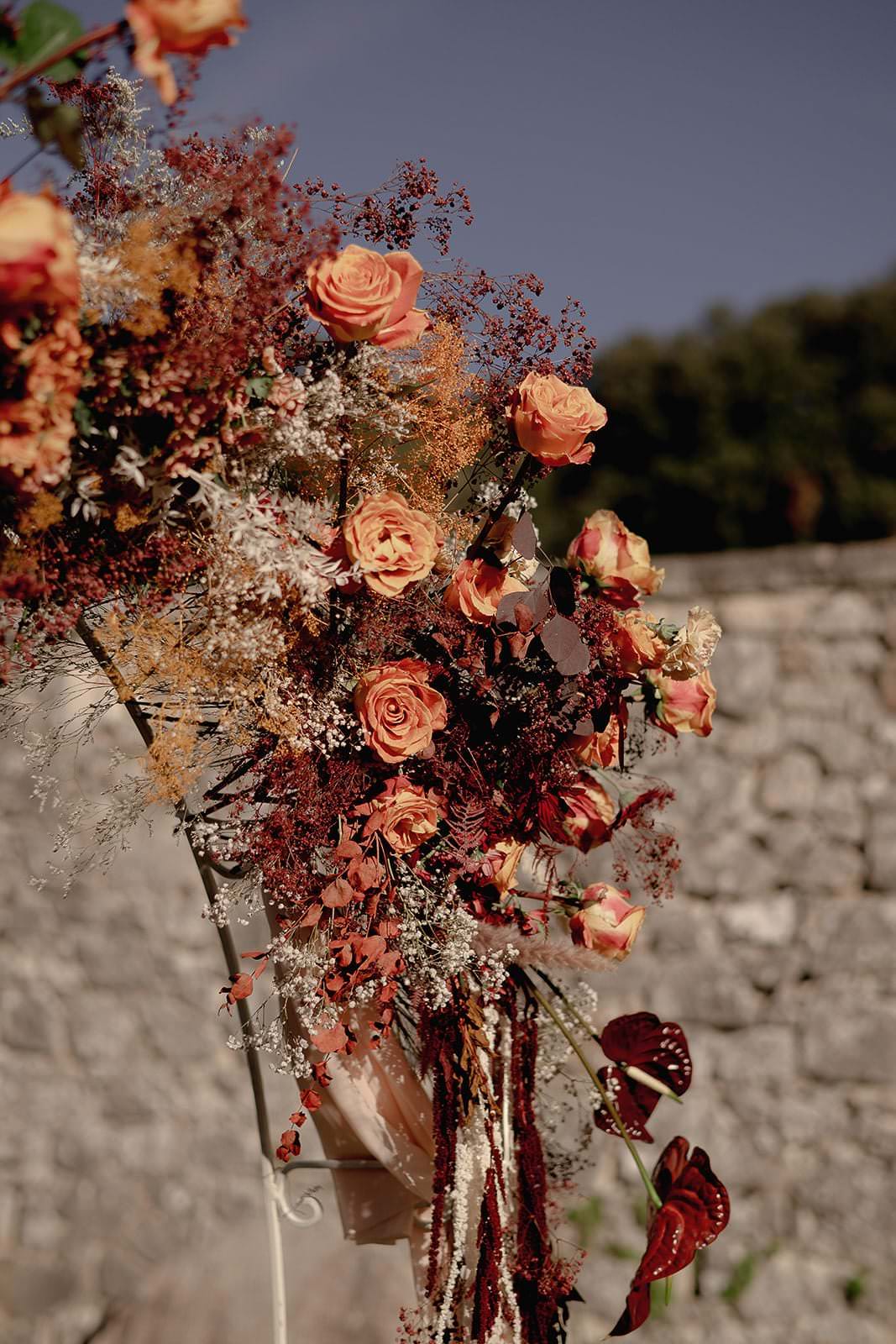 A vibrant floral arrangement featuring orange and peach roses, surrounded by dried foliage in reds and browns. Set against a stone wall under a clear blue Tuscan sky, this exclusive wedding display by Federico Pannacci boasts a cascading design, adding elegance to the breathtaking scene. Tuscan Wedding Photographer