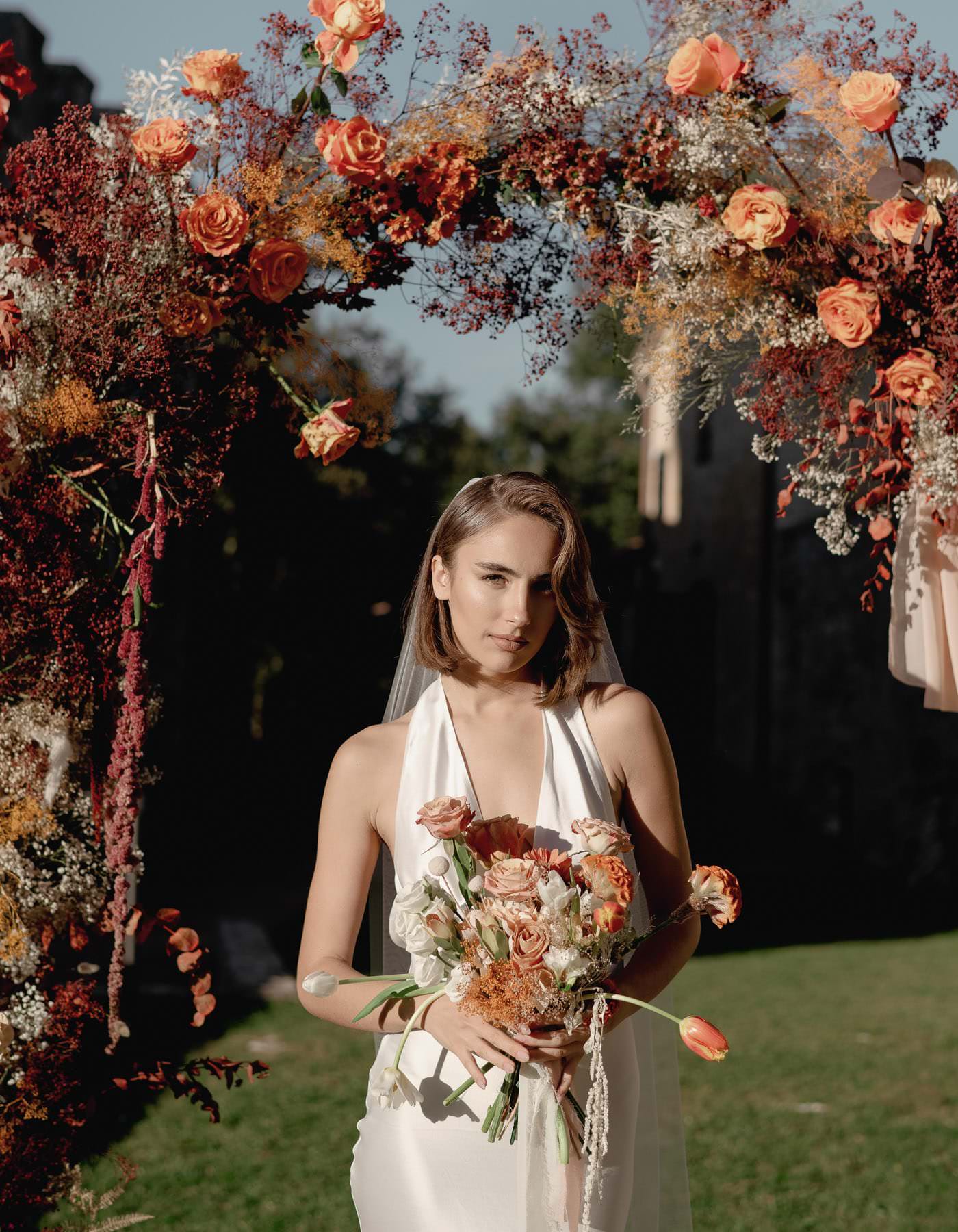 A woman in a white dress stands beneath a floral archway at the enchanting Palazzo Al Piano Wedding. The arch is adorned with red, orange, and white flowers, matching her bouquet. Sunlight casts a natural glow over the serene scene, with lush greenery providing an idyllic backdrop. Tuscan Wedding Photographer