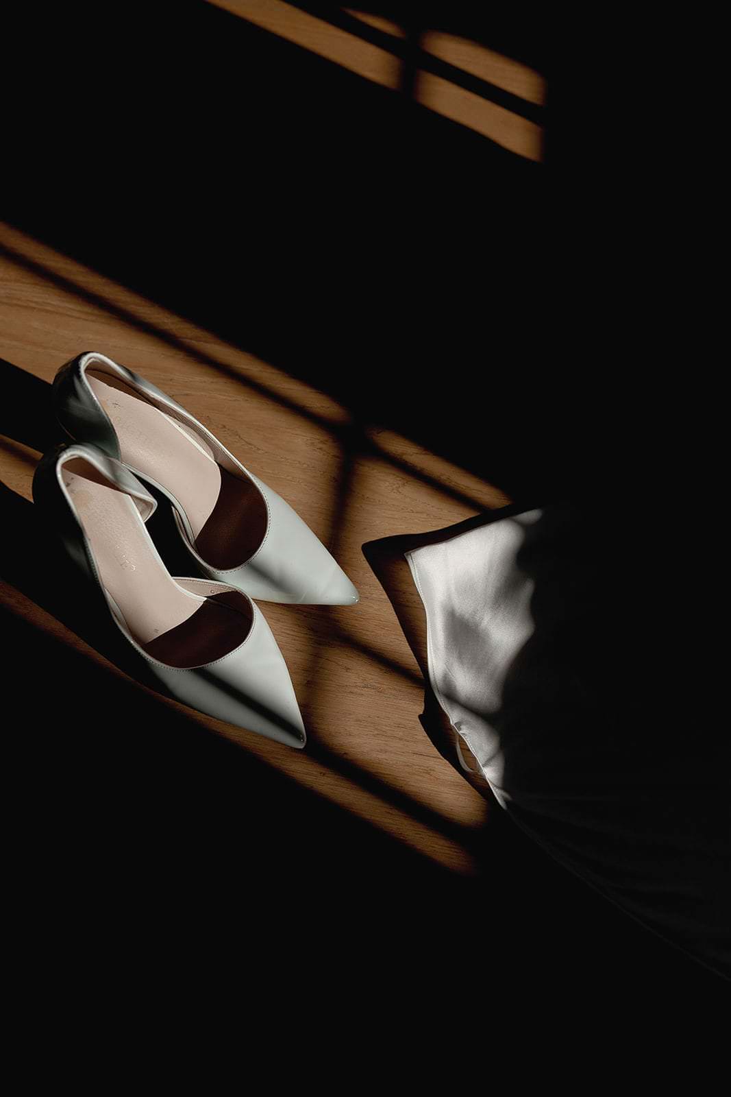 A pair of elegant, light-colored pointed high heels rest on a wooden floor at a Palazzo Al Piano wedding. Sunlight filters through a window, casting geometric shadows. Part of a white fabric, possibly a dress, is visible in the lower right corner, captured beautifully by Federico Pannacci Photography. Tuscan Wedding Photographer