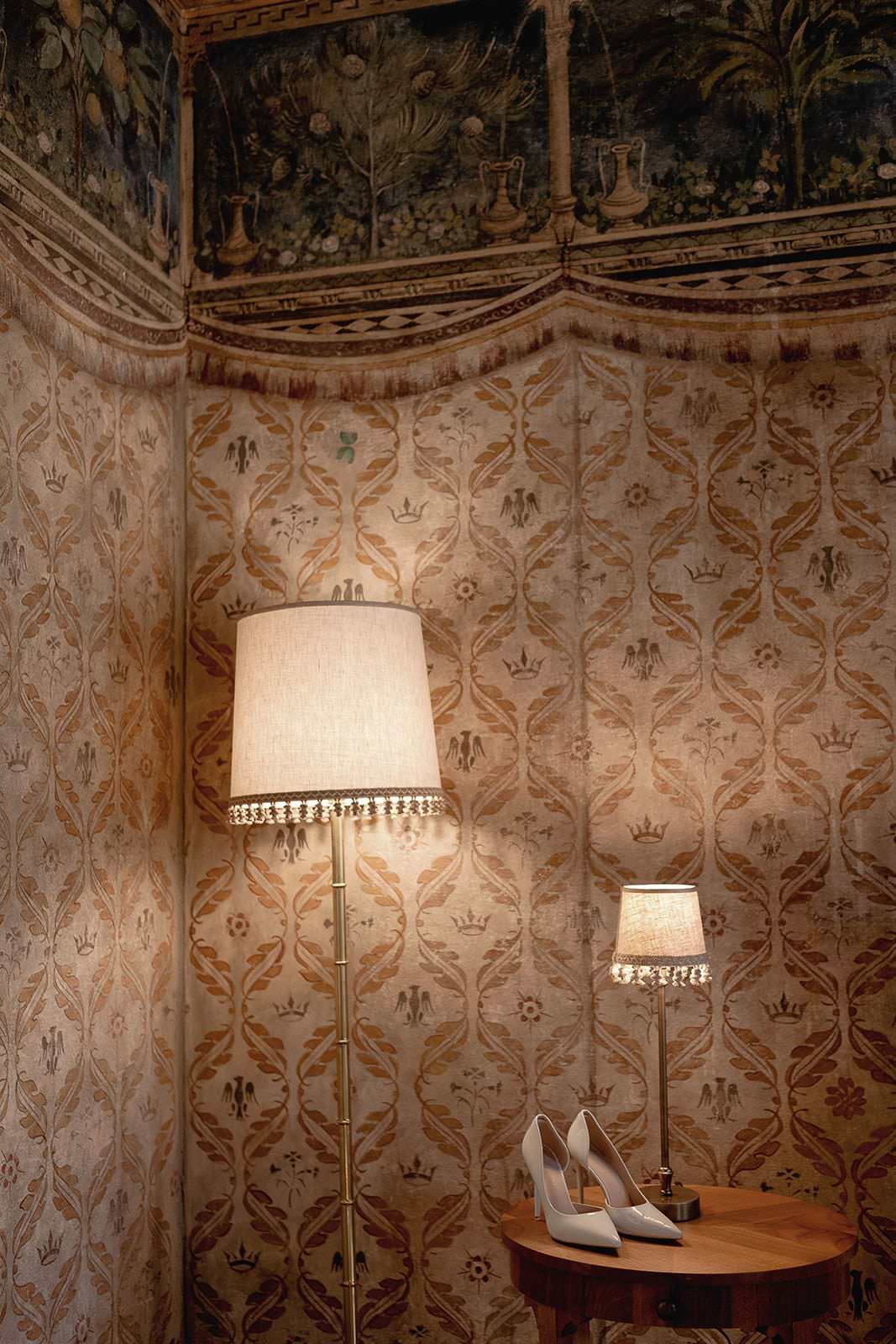 A warmly lit room features ornate wallpaper with floral patterns. A tall floor lamp and a matching table lamp cast a soft glow, reminiscent of a Palazzo Al Piano wedding in Tuscany. On a wooden side table beneath the lamp are elegant white high-heeled shoes, adding sophistication to the cozy setting. Tuscan Wedding Photographer