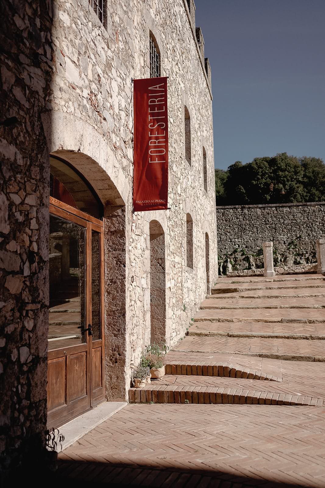 A stone building with arched wooden doors graces the sunlit, brick-paved courtyard of Palazzo Al Piano. A vertical red banner reading Foresteria hangs proudly. The clear blue sky and a stone wall adorned with greenery create the perfect backdrop for a Tuscany wedding, captured by Federico Pannacci Photography. Tuscan Wedding Photographer