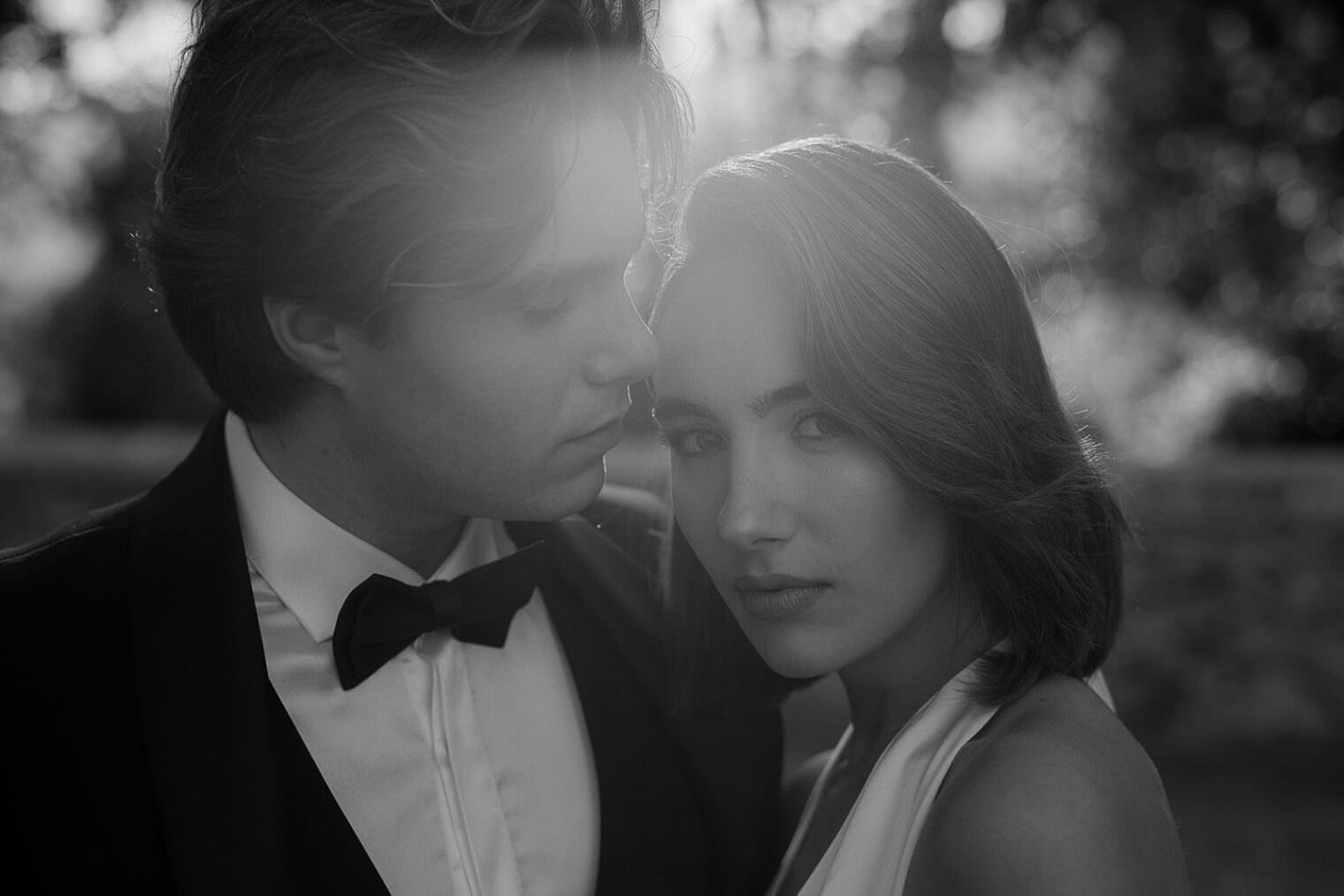 A black and white photo captures a couple in an intimate, romantic atmosphere. The man, in a tuxedo and bow tie, gazes softly at the woman. She wears a Grace Loves Lace Suki dress, short hair gently framing her face as she looks directly at the camera. Its like a timeless wedding in Tuscany. Tuscan Wedding Photographer