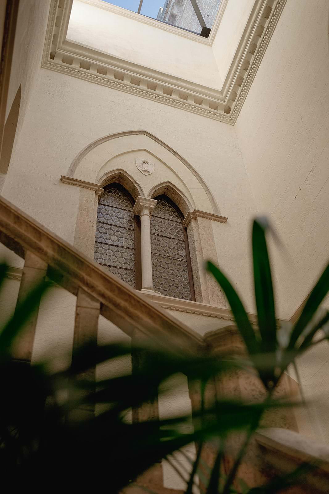 A vintage stone staircase ascends towards a pointed, arched window with intricate, circular glass panels. Sunlight filters in from a skylight above, casting a warm glow perfect for a Palazzo Al Piano wedding in Tuscany. A green plants leaves partially frame the foreground, adding a touch of nature. Tuscan Wedding Photographer