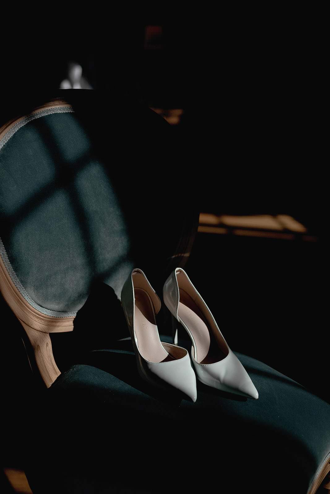 A pair of elegant white high-heeled shoes sit on a dark green vintage chair, captured in an ethereal moment by Federico Pannacci Photography. Soft sunlight filters through the window, casting grid-like shadows across the fabric, echoing the timeless romance of a Palazzo Al Piano wedding in Tuscany. Tuscan Wedding Photographer