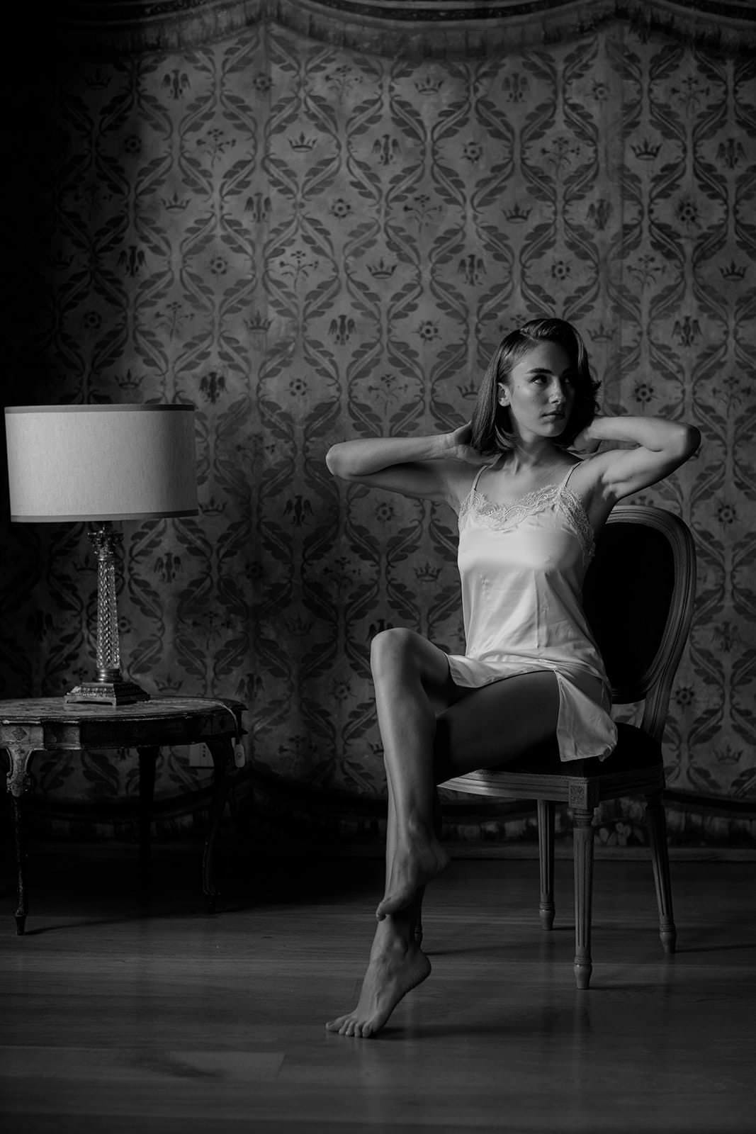 A woman in a silky Grace Loves Lace Suki Dress sits elegantly on a chair in a dimly lit room. She holds her hands behind her head, legs crossed. A table with a lamp is beside her, while the patterned wall enhances the vintage atmosphere of this black-and-white scene. Tuscan Wedding Photographer