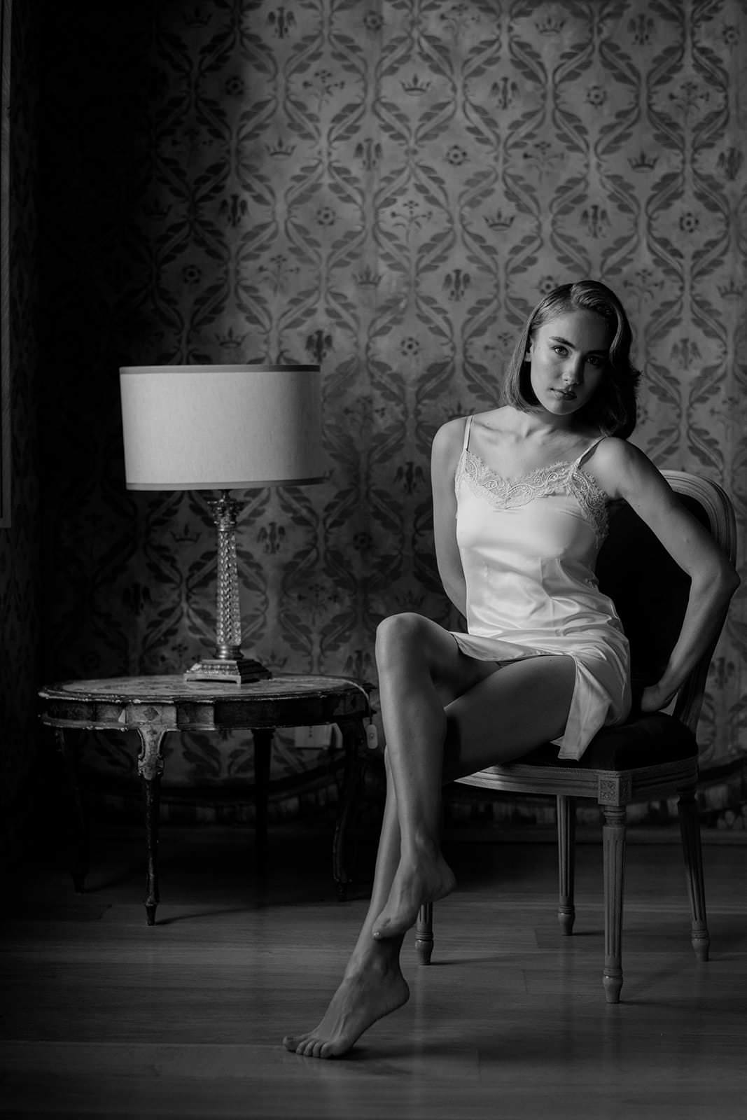A woman in a Grace Loves Lace Suki dress sits on a chair in a dimly lit room with patterned wallpaper. She is barefoot, posing elegantly. To her left is an ornate side table with a lamp. The scene, captured in black and white, exudes the vintage sophistication of a wedding in Tuscany. Tuscan Wedding Photographer