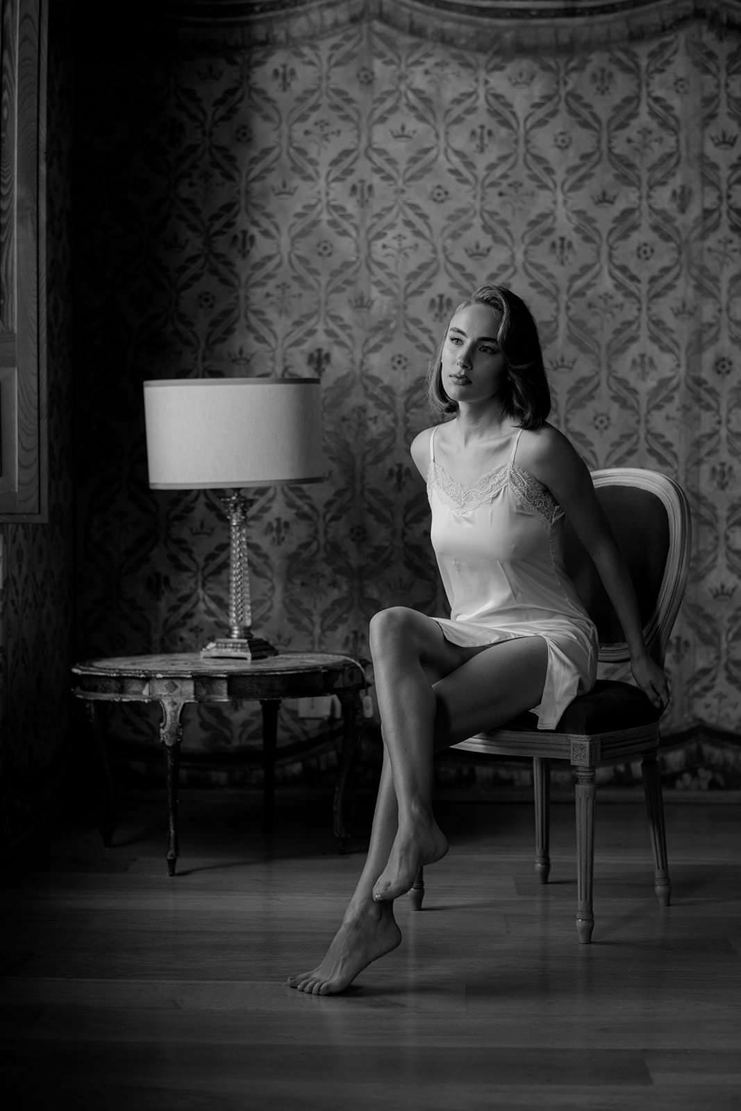 A woman in a Grace Loves Lace Suki dress sits on a chair with her legs crossed, surrounded by ornate wallpaper. Beside her, a round table with a lamp adds to the softly lit ambiance. She gazes thoughtfully toward the window, reminiscent of an elegant wedding in Tuscany captured in black and white. Tuscan Wedding Photographer